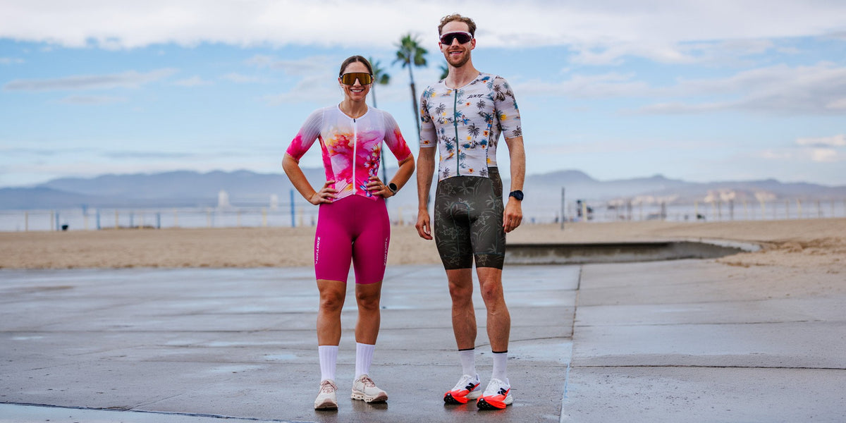 Two cyclists standing on a wet boardwalk by the beach, wearing colorful cycling jerseys and shorts. The woman wears a pink and white jersey with matching pink shorts, and the man wears a blue and white patterned jersey with dark gray shorts.
